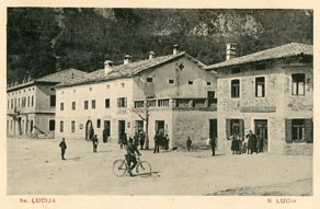 Nekdanje trgovine ob glavnem trgu na Mostu na Soči. V stavbi v sredini je že pred prvo vojno posloval veletrgovec Kovačič, po vojni je njegovo dejavnost nadaljevala hči Marija Alma z možem Eligijem Marego. (B. Mlakar, zasebna last)